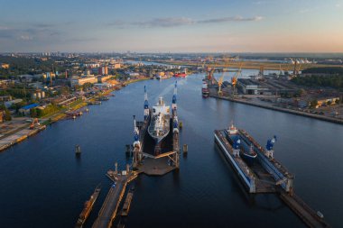Riga, Letonya 'da bir tersane, gün batımında büyük bir gemi kuru bir rıhtımda, endüstriyel vinçler, binalar, ve su yolu kenarında uzak bir şehir manzarası.