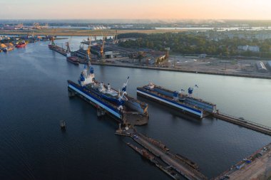 Riga, Letonya 'daki bir tersanede kuru rıhtımlar, vinçler ve sanayi yapılarında büyük gemiler bulunuyor. Günbatımı aydınlatma yeşillik ve kentsel alanları vurguluyor.