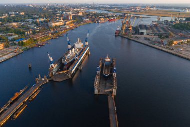 Riga, Letonya 'daki bir tersanede, sıcak gün batımında şehir manzarası olan kuru rıhtımlarda iki gemi, mavi vinçler ve Daugava Nehri yer alıyor..