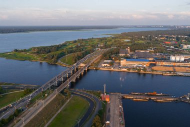 Riga, Letonya 'daki Daugava Nehri' ni geçen çok şeritli bir köprünün hava görüntüsü. Endüstriyel binalar, yeşil alanlar ve akşam ışığında sakin sular..