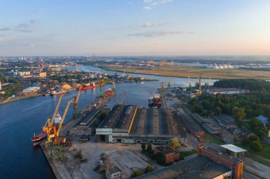 Riga, Letonya 'daki bir tersanenin hava görüntüsünde vinçler, kenetlenmiş gemiler ve Daugava Nehri yer alıyor. Sanayi bölgesini çevreleyen kentsel yapılar ve yeşillikler.