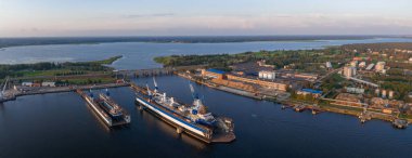 Riga, Letonya 'da geniş bir nehir boyunca vinçleri, gemileri ve endüstriyel tesisleri olan bir tersane. Arka planda yeşil bir manzaraya bir köprü bağlanıyor.