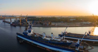 Riga tersanesinin üzerinde canlı bir gün batımı. Mavi ve beyaz gemiler kenetlenmiş, rıhtım boyunca vinçler ve arka planda kentsel yapılar var..