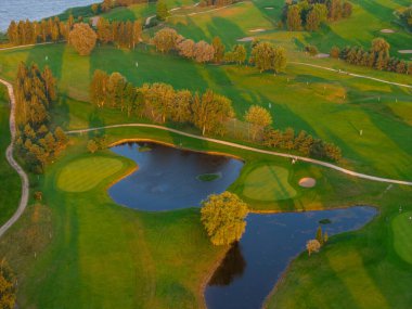 Gün batımında yemyeşil bir golf sahası. Çim yollarının, kum sığınaklarının ve su tehlikelerinin olduğu. Bir dolambaçlı yol ve ağaçlar derinlik ekler, arka planda su vardır..