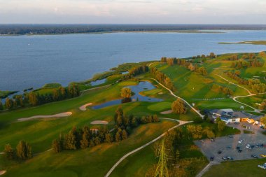 Gün batımında golf sahasının manzaralı manzarası, kum sığınakları, su tehlikeleri ve büyük bir su kütlesi yakınında bir kulüp evi..