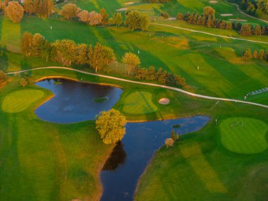 Gün batımında bir golf sahasının canlı hava görüntüsü, yeşil çimenler, kum sığınakları, bir su tehlikesi, ve sakin bir manzarada golfçüler.
