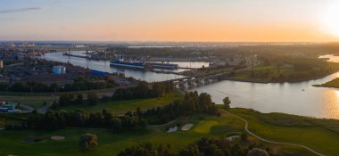 Riga, Letonya üzerinde sakin bir gün batımı, bir nehir, bir köprü, rıhtımlı tekneleri olan bir tersane, yemyeşil bir arazi ve görünür çim yolları olan bir golf sahası.