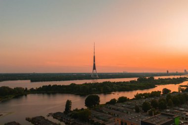 Riga TV Kulesi, yeşil, Daugava Nehri ve ön planda bir yerleşim alanı ile çevrili turuncu ve pembe bir günbatımı gökyüzüne karşı dimdik ayakta duruyor..