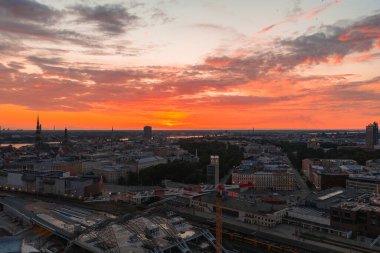 Riga Eski Kasabası 'nın kuleleri, Aziz Peter Kilisesi de dahil, gün batımında Daugava Nehri' nin yanında duruyor. Kısmen inşa edilmiş bir yapı modern bir karşıtlık ekler.