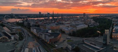 Riga, Letonya, gün batımında Eski Şehir kuleleri, Riga TV Kulesi, Ulusal Kütüphane, demiryolu rayları ve Daugava Nehri üzerindeki yansımalarla.