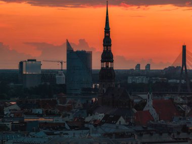 St. Peter Kilisesi 'nin silueti, Riga, Letonya' da günbatımında, eski kasaba çatıları, Vansu Köprüsü ve arka planda modern binalar ile aydınlanıyor..