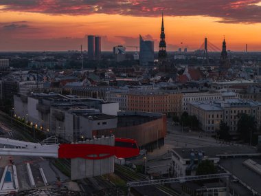 Gün batımında Riga şehrinde Aziz Peter Kilisesi kulesi, Vansu Köprüsü, yüksek raylarda kırmızı ve beyaz bir tren ve tarihi ve modern binalar bulunmaktadır..