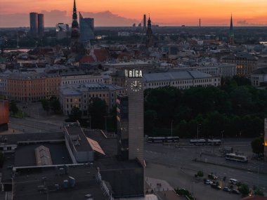 Riga, Letonya ufuk çizgisi günbatımında modern bir saat kulesine sahip 'RIGA' aydınlatılmış, tarihi kuleler St. Peter Kilisesi gibi, ve kentsel yeşil alanlar.
