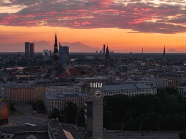 Riga 'nın St. Peter Kilisesi ve Riga Katedrali de dahil olmak üzere eski şehir kuleleri, ön planda saat kulesi bulunan canlı turuncu ve pembe bir günbatımına karşı durmaktadır..