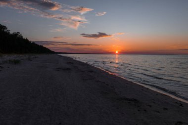 Letonya, Jurmala 'da kumlu bir plajın üzerinde ılık turuncu bir parıltı olan sakin bir günbatımı sahnesi. Sakin deniz, yumuşak bulutlarla çizilmiş gökyüzünü yansıtır..