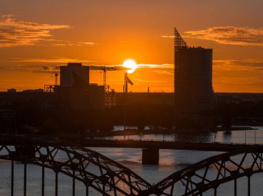 Riga TV Kulesi ve şehir silueti gün batımında, Daugava Nehri ve ön planda bir köprü ile göze çarpıyor. Merkezin yakınında bir Letonya bayrağı görülüyor.
