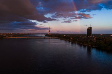 Riga, Letonya manzarası, Riga TV Kulesi, soluk bir gökkuşağı, Daugava Nehri, bir köprü, modern binalar ve dramatik günbatımı bulutları..