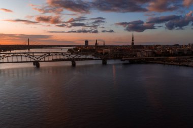 Riga, Letonya, gün batımında St. Peter Kilisesi kulesi, tarihi eski kasaba binaları, Vansu Köprüsü ve sıcak turuncu gökyüzünün altındaki Daugava Nehri..