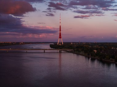 Riga, Letonya 'daki Daugava Nehri üzerinde sakin bir gün batımı, kırmızı ve beyaz Riga TV Kulesi, bir köprü, yumuşak pembe ve mor gökyüzü.