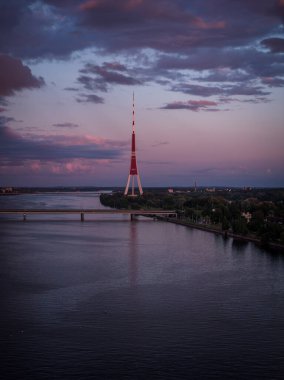 Riga TV Kulesi, gün batımında Daugava Nehri 'nin kıyısında, denizi geçen bir köprüyle ayakta duruyor. Gökyüzünün pembe ve mor renkleri sakin nehri yansıtıyor..