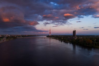 Riga, Letonya 'daki Daugava Nehri manzarası, Riga TV Kulesi, modern bir gökdelen, yemyeşil ve renkli günbatımı yansımaları yer alıyor..