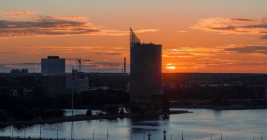 Riga, Letonya 'nın gün batımında üçgen çatısı, Daugava Nehri yansımaları, ağaçlar, bayrak direği ve kentsel kalkınma vinçleriyle yüksek bir gökdelenin görüntüsü.