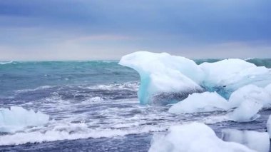 Turkuaz buzdağları, dalgalar çarpıp hareket oluştururken siyah kumların üzerinde dinlenir. Deniz kuşları, sakin bir gökyüzü ve derin mavi bir okyanusun altında görünür..