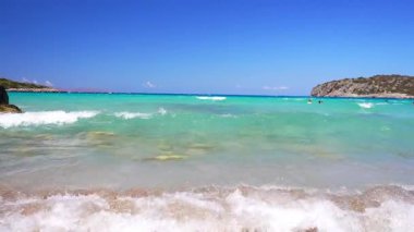 Turkuaz dalgalar Yunanistan 'ın Girit kentindeki kumlu bir kıyıya vururken, yüzücüler berrak sularda yüzüyor. Rocky Hills kıyı şeridini parlak mavi bir gökyüzünün altına kurar..