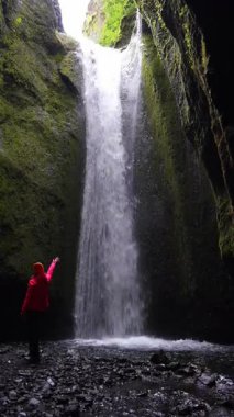 Bir şelale yosun kaplı bir uçurumdan İzlanda 'daki kayalık bir havuza akar. Kırmızı ceketli biri tabanın yanında yürüyor, ölçü ve hareket ekliyor..