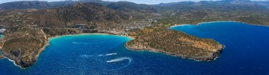 Yunanistan 'ın Girit kentindeki Voulisma Sahili' nin hava manzarasında turkuaz sular, hilal şeklinde bir kıyı şeridi, kayalık bir arazi ve arka planda küçük bir kasaba görülüyor..