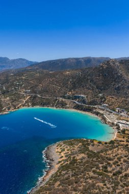 Yunanistan 'ın Girit kentindeki Voulisma Sahili' nin hava manzarasında derin mavi sulara turkuaz, hilal kıyı şeridi, engebeli tepeler ve bir tekne yolu yer alıyor..