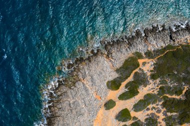 Girit 'teki Voulisma Sahili yakınlarında canlı mavi suları, yeşil bitki örtüsü ve turuncu kahverengi toprağı olan kayalık bir sahil manzarası..