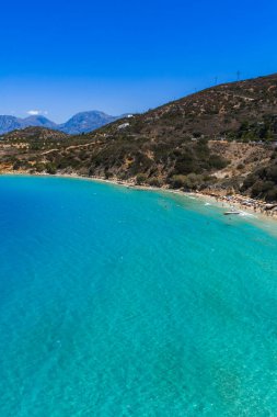 Yunanistan 'ın Girit kentindeki Voulisma Sahili' nin hava manzarasında turkuaz sular, hilal şeklinde kıyı şeridi, engebeli tepeler ve mavi gökyüzünün altındaki uzak dağlar yer alıyor..