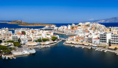 Girit 'teki Agios Nikolaos' un hava manzarası, teknelerin, beyaz ve pastel binaların, dar bir kanalın, küçük bir adanın ve uzak dağların bulunduğu bir limanı gösteriyor..