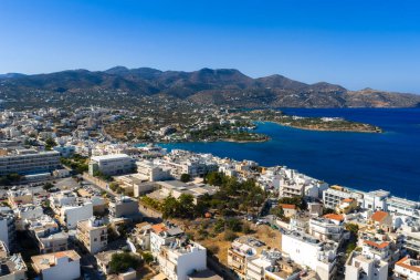 Girit 'teki Agios Nikolaos' un hava manzarasında Akdeniz kıyısındaki beyaz ve pastel binalar, küçük bir yarımada ve arka planda engebeli dağlar görülüyor..