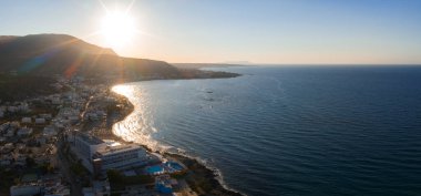 Stalida, Girit 'in hava manzarası, günbatımında bir kıyı kasabasını gösteriyor. Kıvrımlı bir koy, binalar, havuz ve arka planda Ege Denizi var..