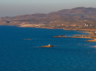 Girit, Stalida 'nın havadan görünüşü, beyaz bir yapısı, kumlu plajları, sahil binaları ve altın saat ışığı altında engebeli tepeleri olan kayalık bir adacığı gösteriyor..