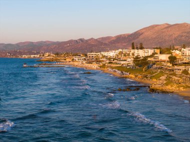 Stalida, Girit 'in havadan görünüşü, arka planda şemsiyeli kumlu sahilleri, turkuaz suları, kıyı binaları ve güneşli dağları gösteriyor..