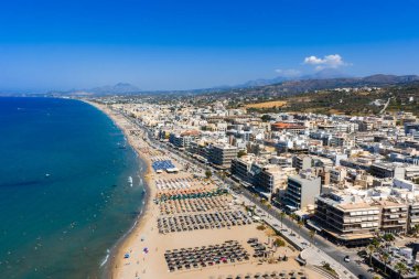Yunanistan 'ın Girit kentindeki Rethymno kentinin hava manzarasında, içinde şemsiyeler, turkuaz deniz, kümelenmiş binalar ve dağ arkaplanı bulunan kumlu bir plaj yer alıyor..
