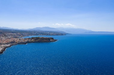 Yunanistan 'ın Girit Adası' nın havadan görünüşü kayalık bir kıyı şeridi, canlı bir mavi deniz ve berrak mavi gökyüzünün altındaki dağlar gösteriyor..