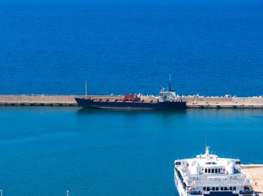 Girit, Rethymno 'da bir liman manzarası, rıhtımda bir kargo gemisi, beyaz bir feribot, beton bir iskele, ve güneşli gökyüzünün altında sakin mavi deniz..