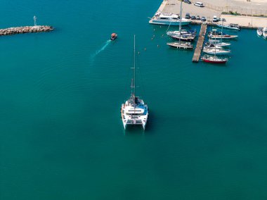 Girit, Rethymno 'da bir marinanın havadan görüntüsü, turkuaz sularda katamaran, demirlemiş tekneler, bir motorlu tekne, kayalık bir dalgakıran ve park halindeki arabalar..