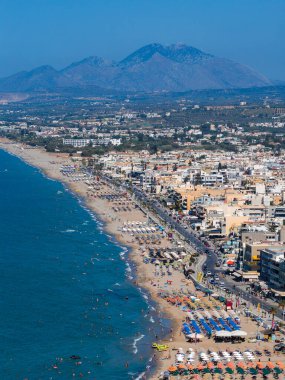 Yunanistan 'ın Girit kentindeki Rethymno kentinin havadan görünüşü, şemsiyeli kumlu bir sahil, hareketli bir gezinti güvertesi, modern ve geleneksel binalar ve dağları gösteriyor.