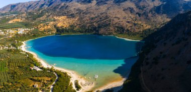 Yunanistan 'ın Girit kentindeki Kournas Gölü' nün hava manzarasında turkuaz sular, tekneleri, engebeli tepeleri, zeytinlikleri ve dağınık binaları gösteren kumlu bir sahil görülüyor..