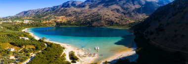 Yunanistan 'ın Girit kentindeki Kournas Gölü' nün havadan görünüşü, turkuaz suları, şemsiyeli kumlu bir kıyı şeridi, tekneler, zeytinlikler ve engebeli dağları gösteriyor.