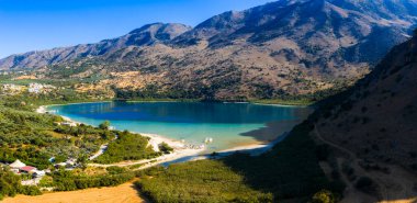 Yunanistan 'ın Girit kentindeki Kournas Gölü' nün hava manzarası, turkuvaz suları, yemyeşil yemyeşil, kumlu kıyılar, küçük kayıklar ve arka planda engebeli dağlar gösteriyor.