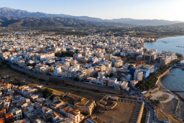 Günbatımında yukarıdan görülen Chania, Girit 'te beyaz ve pastel binalar, bir Venedik duvarı, berrak mavi sular ve uzakta engebeli dağlar bulunur..