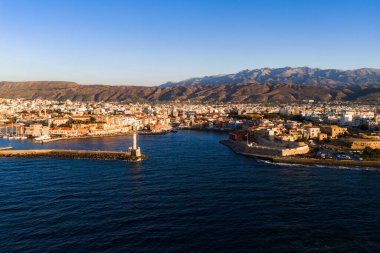 Sıcak günbatımında Venedik Limanı, ikonik deniz feneri, tarihi binalar ve dağların yer aldığı Chania, Girit, Yunanistan 'ın hava manzarası.