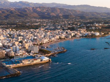 Günbatımında Girit, Yunanistan 'daki Chania' nın hava manzarası. Beyaz ve pastel binalar, rıhtımlar, tekneler, turkuaz sular ve görünür dağlarla kıyı şeridi boyunca uzanır..