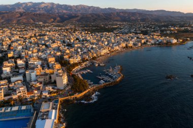 Chania, Girit, Yunanistan kıyı binalarını, tekneli bir marinayı, günbatımını yansıtan sakin denizi ve arka planda engebeli dağları gösteren hava manzarası.